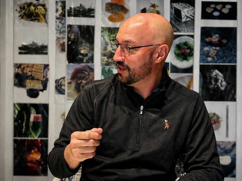 Bald man with glasses and a beard wearing a dark zip sweater, gesturing with his right hand in front of a photo-covered wall behind him.