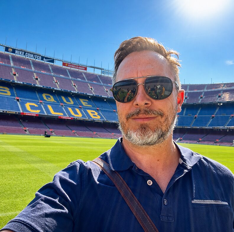 Selfie of a man wearing sunglasses at a sunny football stadium, with a green field and blue and red seats behind; yellow letters spell 'QUE CLUB' in the stands.